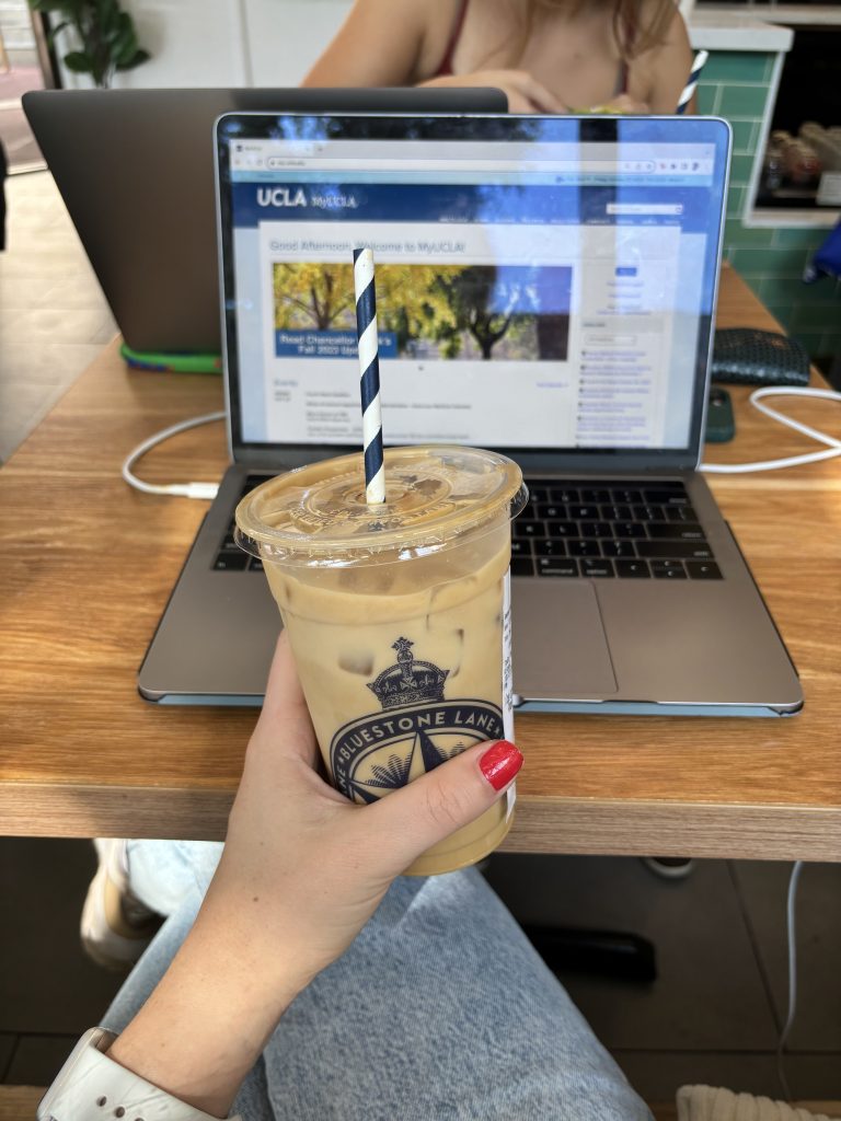 A person holds up a Bluestone Lane coffee cup in front of their computer screen. Students often go to this coffee shop to sit down and get work done. Photo by Veronika Znam/BruinLife