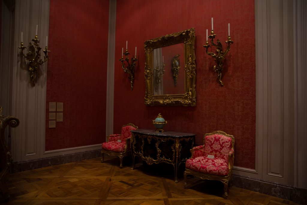 A bold display of furniture is displayed in a corner, with two red chairs, a small cabinet, two candelabras and a mirror. Photo by Katelyn Michel/BruinLife.