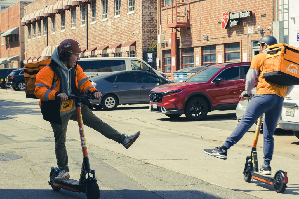 Aidan Seidle kicks off a lively ride on an electric scooter, his face alight with the thrill of motion and the open road ahead. Meanwhile, his colleague zooms ahead, fully immersed in the hustle of the urban delivery life. Photographed by Eliza Loventhal/BruinLife.