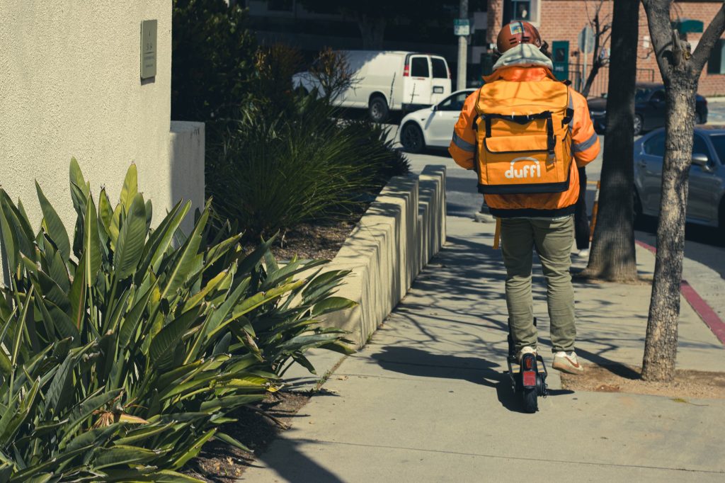 A Duffl employee, recognizable by the signature bright orange backpack, cruises down a Westwood sidewalk on an electric scooter, swiftly navigating towards the next delivery destination. Photographed by Eliza Loventhal/BruinLife.