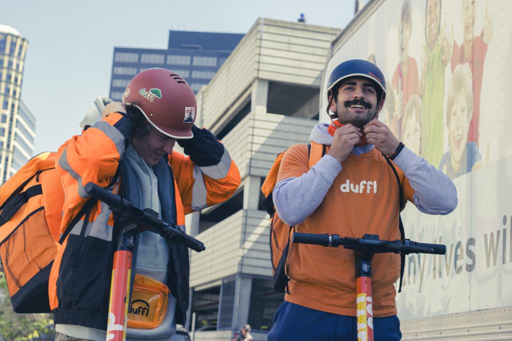 Safety and smiles go hand in hand as Aidan Seidle and co-worker gear up for the road, fastening their helmets before they embark on Westwood' streets. Their bright attire and easy camaraderie shine against the backdrop of Westwoods landscape, ready for the day's work adventures. Photographed by Eliza Loventhal/BruinLife.
