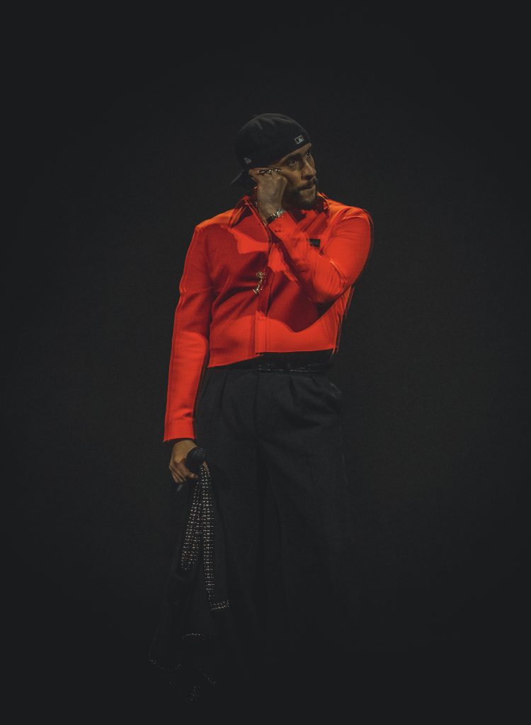 Bad Bunny looks to the right with his hand up to his face. As a critically acclaimed artist with nine Grammy nominations and three wins, Bad Bunny has solidified his place in the music industry and continues to deliver performances that impress beyond expectations.