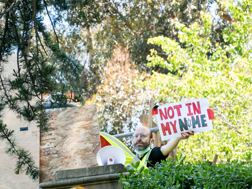 A pro-Palestine organizer appears from behind the encampment walls holding up a sign that reads "NOT IN MY NAME," with the "A" being written with the Star of David. The encampment spread overnight past the Shapiro Fountain, barricading Janss/Kuruvungna Steps and adjacent walkways. Photographed by Finn Martin/Bruinlife.