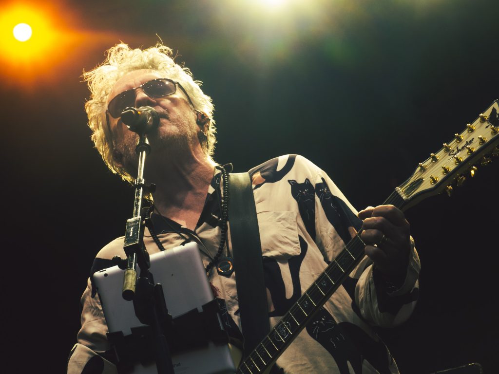 Lead singer Wayne Hussey of The Mission U.K. nears the microphone. An orange and white rail lights glow down from above. Photographed by Finn Martin/BruinLife