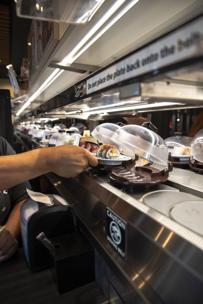 A patron grabs a red dragon roll from the plastic domes as they circulate around the conveyor belt. Photographed by Emily Chandler/BruinLife.