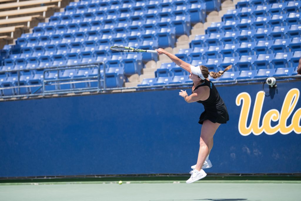 UCLA player swings racket in mid air to serve ball over net.