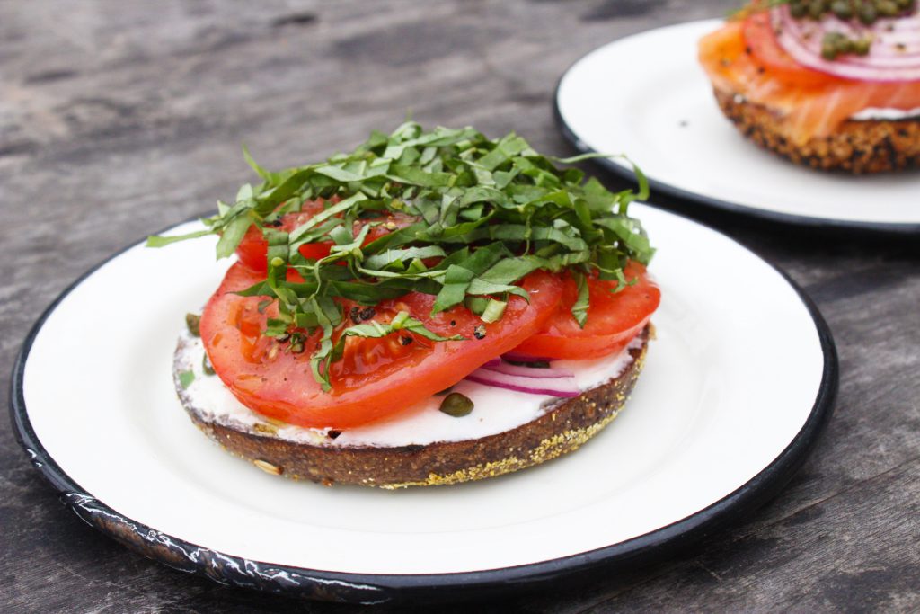 The bottom half of a pumpernickel bagel is topped with two too many tomatoes, a polit mound of chiffonade basil, capers and red onions.