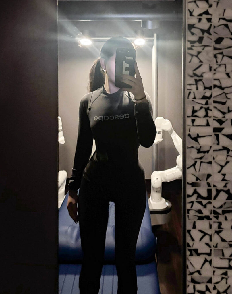 I’m wearing the dark grey Aerwear outfit, a fitted long-sleeve top with matching leggings tucked in, as I take a mirror selfie inside the Aescape massage room. Behind me, the white robotic arms and the blue Aescape treatment table are visible. Photographed by Ellie Mitola/BruinLife.