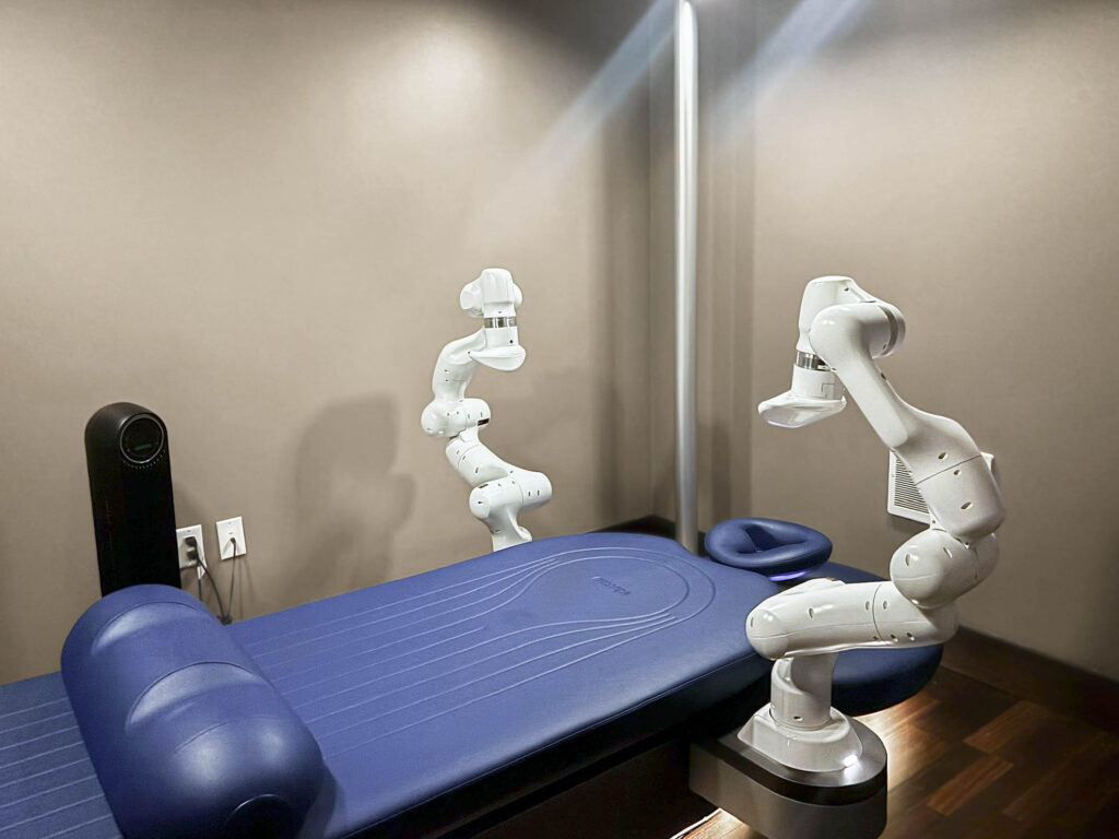 A massage room inside the Equinox Westwood spa featuring the Aescape massage table. The machine has two white mechanical arms positioned above the table that perform the automated massage. The photo shows the table as it appears when you first walk into the room. Photographed by Ellie Mitola/BruinLife.